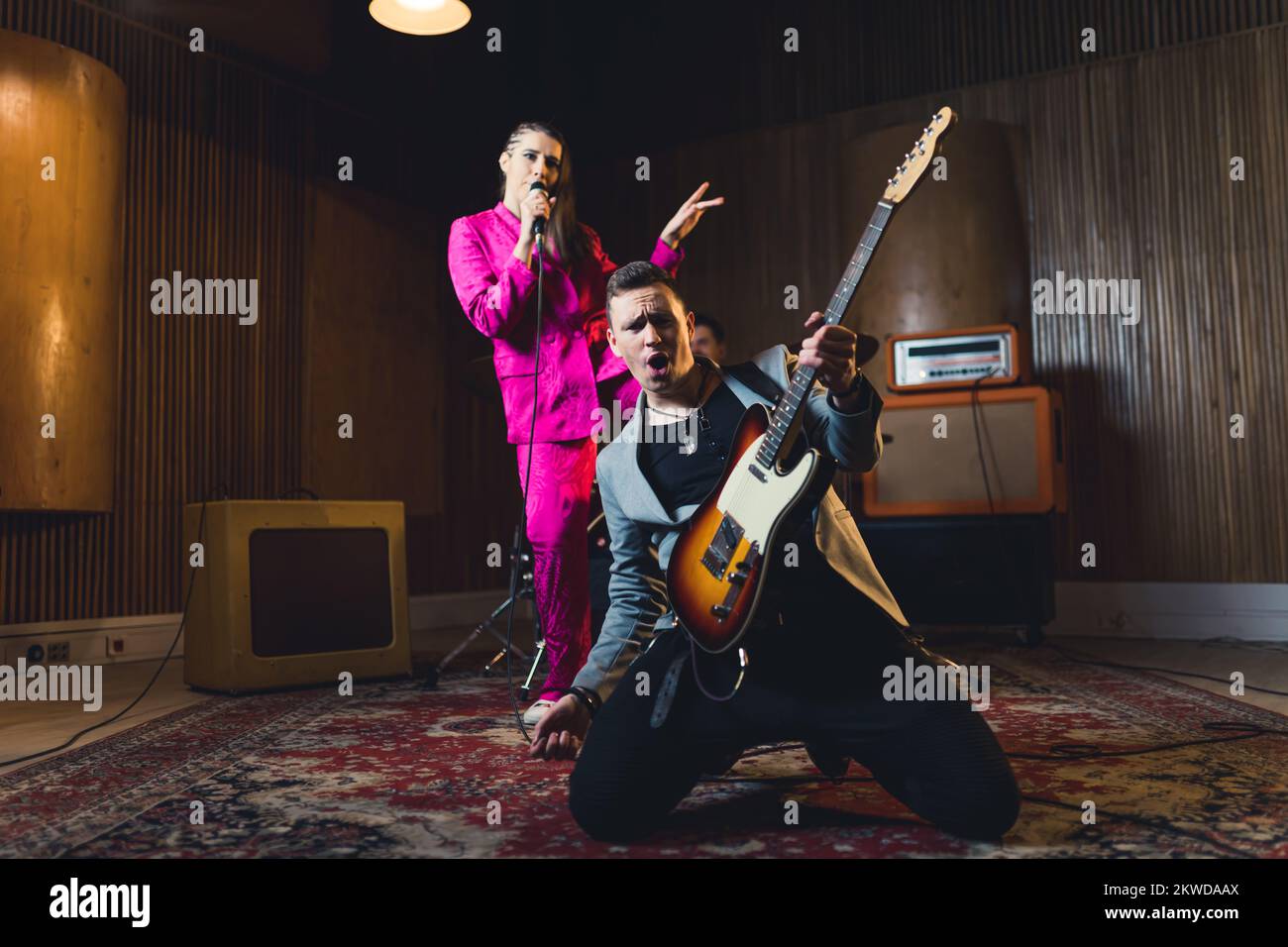 Innenaufnahme eines weißen Mannes mit elektrischer Gitarre, die vor einer Sängerin seiner Band auf dem Teppich kniet. Aufnahmestudio. Hochwertiges Foto Stockfoto