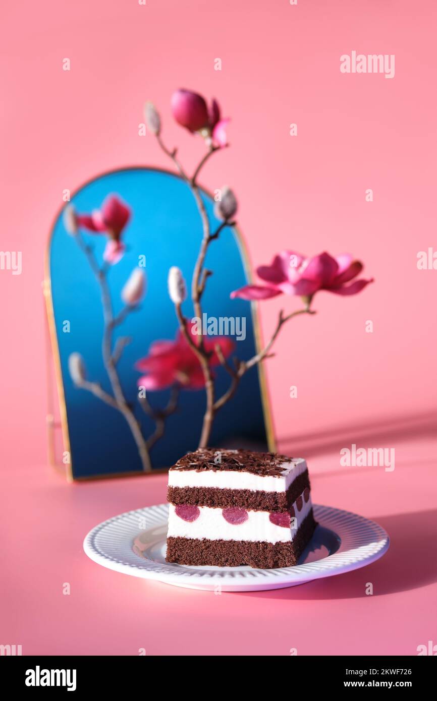 Schokoladenkuchen mit Sauerkirschen. Ein Stück Kuchen auf einem Teller mit Gabel. Süßes Dessert auf pinkfarbenem Hintergrund mit lebhaften Magnolienblüten. Stockfoto