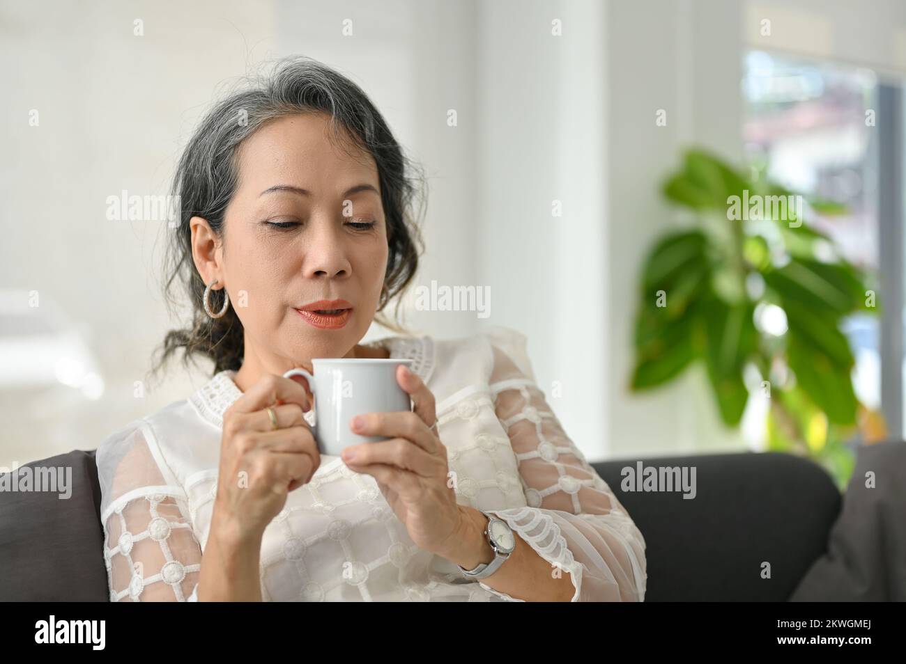 Eine friedliche und entspannte ältere asiatische Frau trinkt gerne morgens Kaffee, während sie in ihrem Wohnzimmer zu Hause chillt. Stockfoto