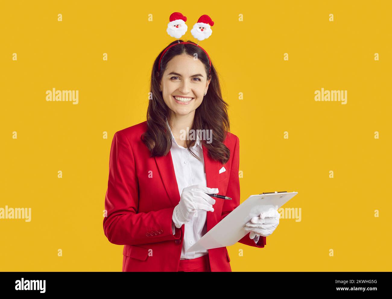 Lächelnde Frau im Weihnachtsmannstil mit Weihnachtsband, das Notizen auf dem Klemmbrett auf gelbem Hintergrund macht. Stockfoto