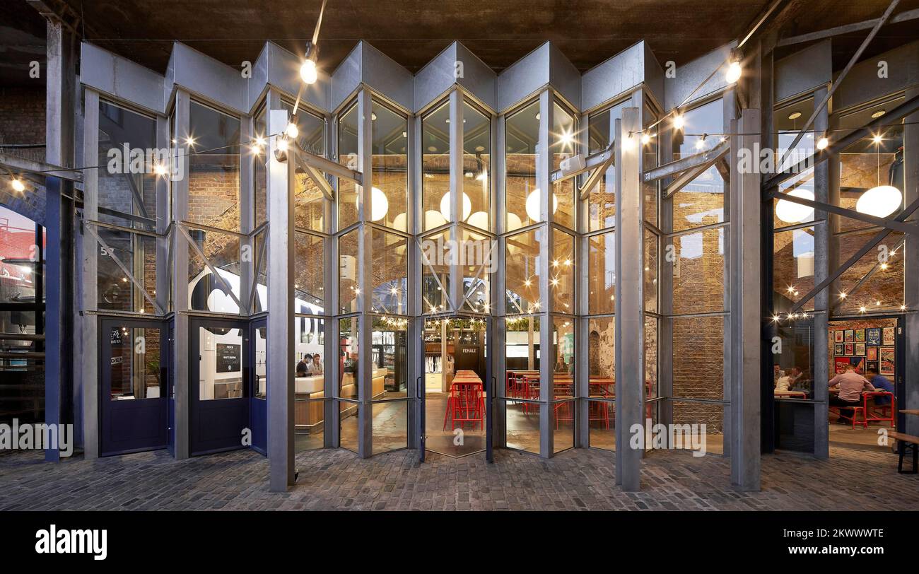 Beleuchtete Innenausstattung in der Abenddämmerung, Blick von Mews. Camden Town Beer Hall, London, Großbritannien. Architekt: Gundry & Ducker, 2021. Stockfoto