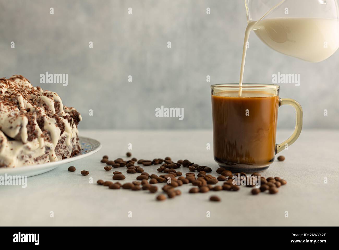 Kaffee mit Creme in einem Glas Becher mit Kaffeebohnen, Cremekuchen. Creme wird aus einem Glaskrug gegossen. Trinken Sie auf hellgrauem Hintergrund. Stockfoto