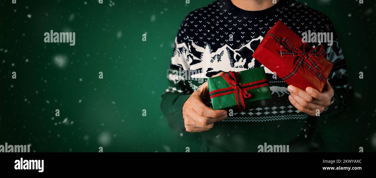 Ein Mann im weihnachtspullover, der Geschenkschachteln in der Hand hält, auf grünem Hintergrund. Banner mit Kopierbereich Stockfoto