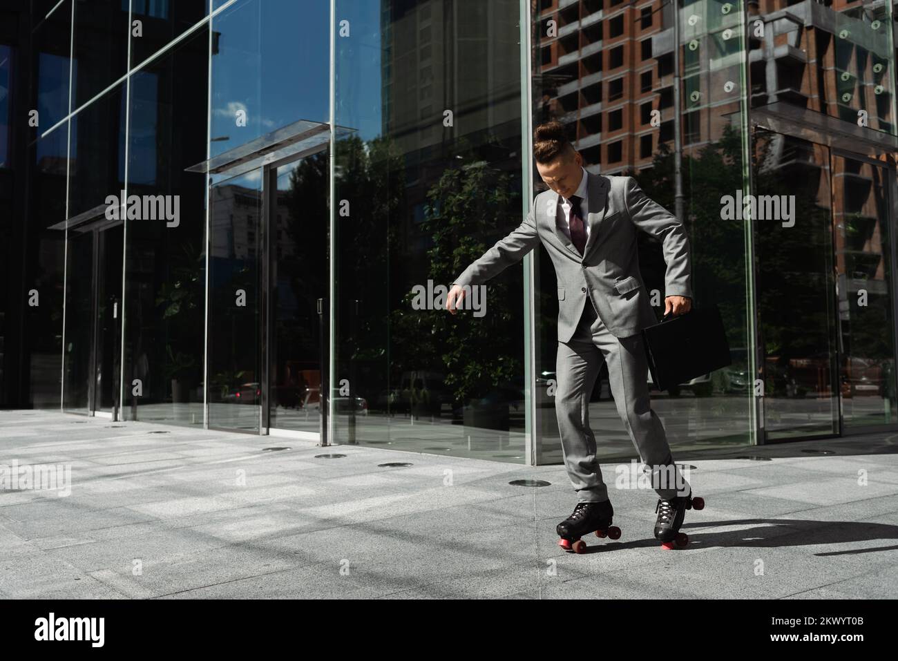 Langer junger Geschäftsmann im grauen Anzug, Rollschuhlaufen in der Nähe eines modernen Gebäudes mit Glasfassade, Stockbild Stockfoto