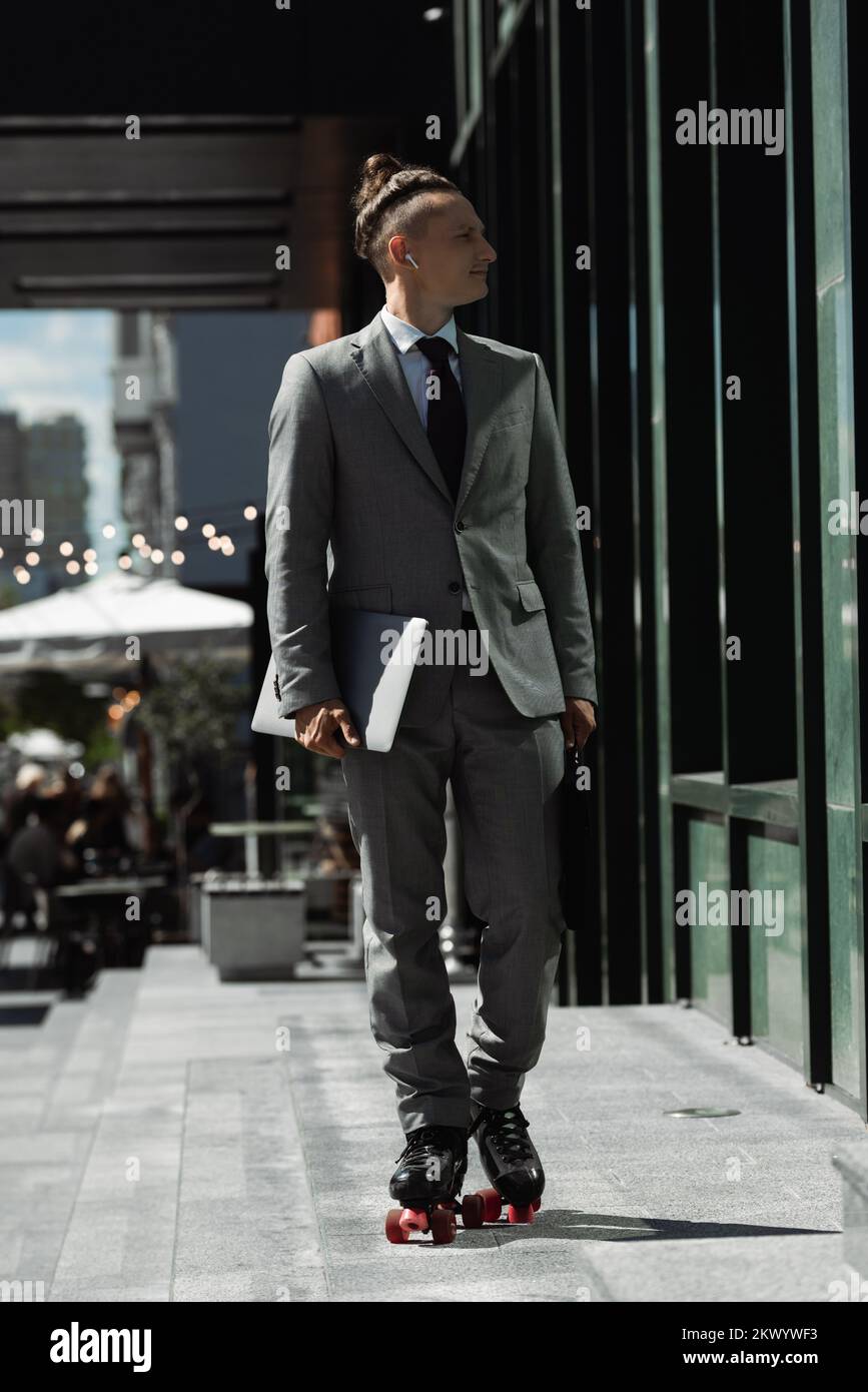 Junger Geschäftsmann im grauen Anzug hält Laptop, während er auf der Straße Rollschuhlaufen geht, Stockbild Stockfoto