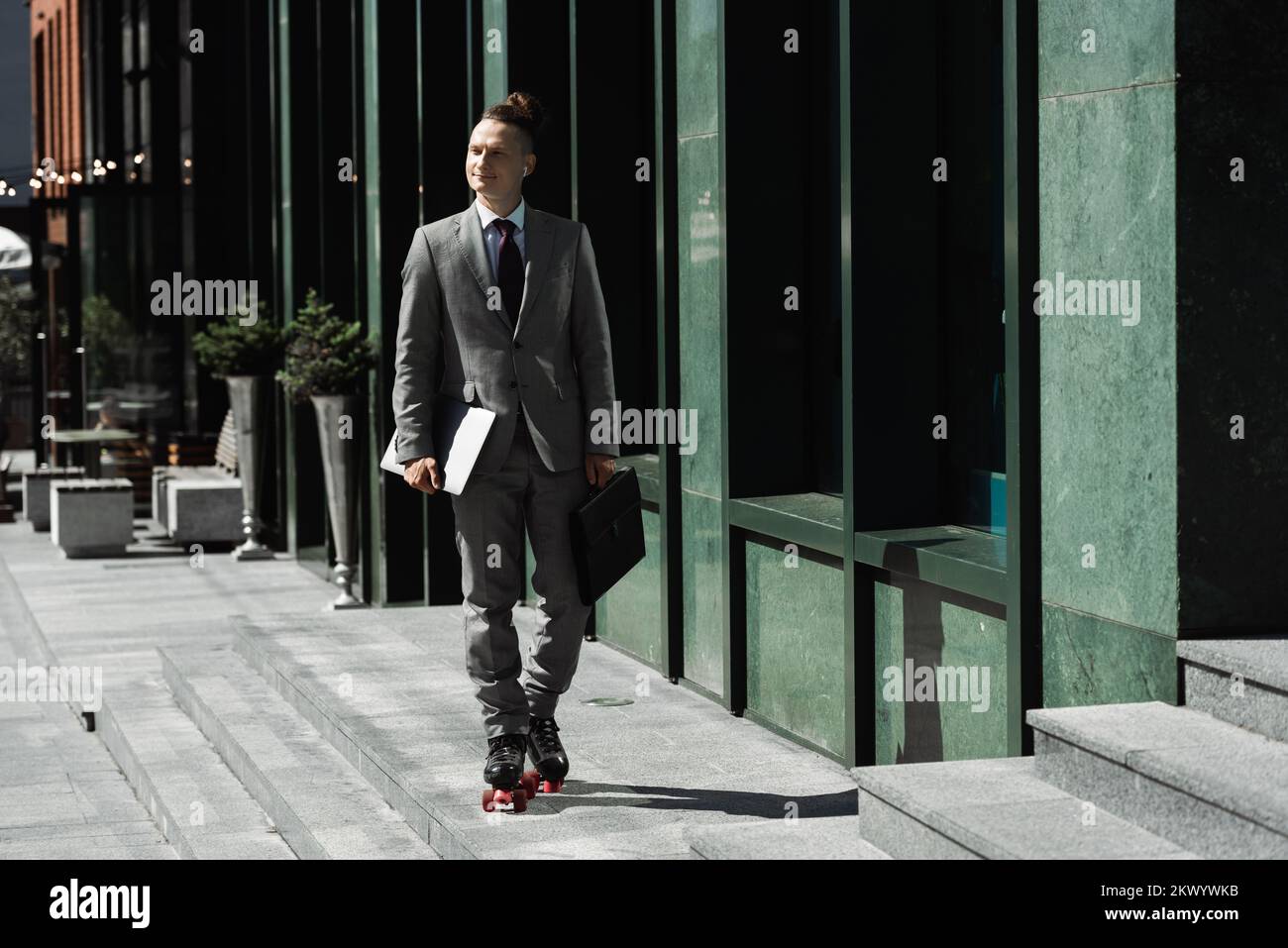Glücklicher Mann in Business-Anzug und Rollschuhen mit Laptop und Aktentasche, während er auf der Straße der Stadt Schlittschuh läuft, Stockbild Stockfoto
