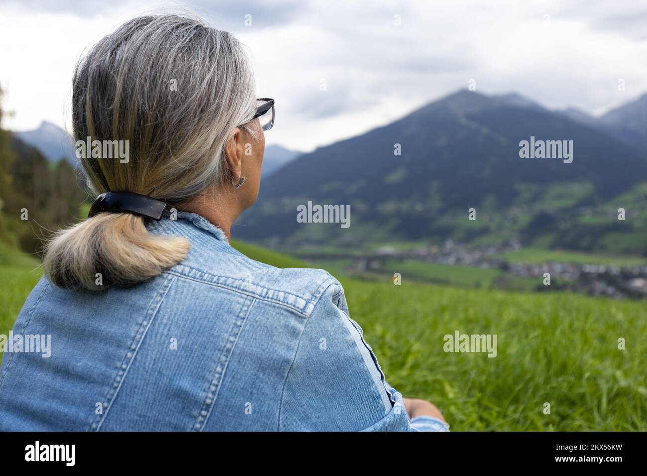 Eine ältere Frau im Jeansanzug sitzt auf einer hohen Wiese und schaut auf die Berge auf der anderen Seite der Schlucht. Stockfoto