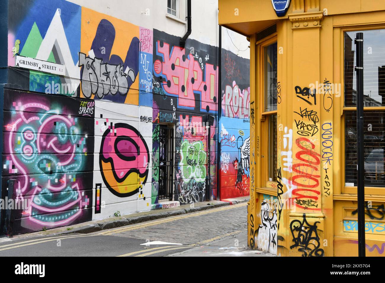 Mit Graffiti und Straßenkunst bedeckte Mauer in Grimsby Street, Shoreditch, London, mit einer gelben Ladenfront mit Graffiti bedeckt Stockfoto