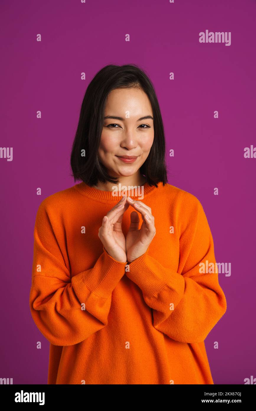 Junge asiatische Frau, die Gesten macht, während sie vor der Kamera lächelt, isoliert über lila Hintergrund Stockfoto