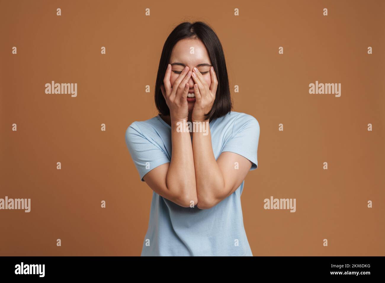 Junge asiatische Frau, die ein T-Shirt trägt, lacht und ihr Gesicht verdeckt, isoliert vor beigem Hintergrund Stockfoto