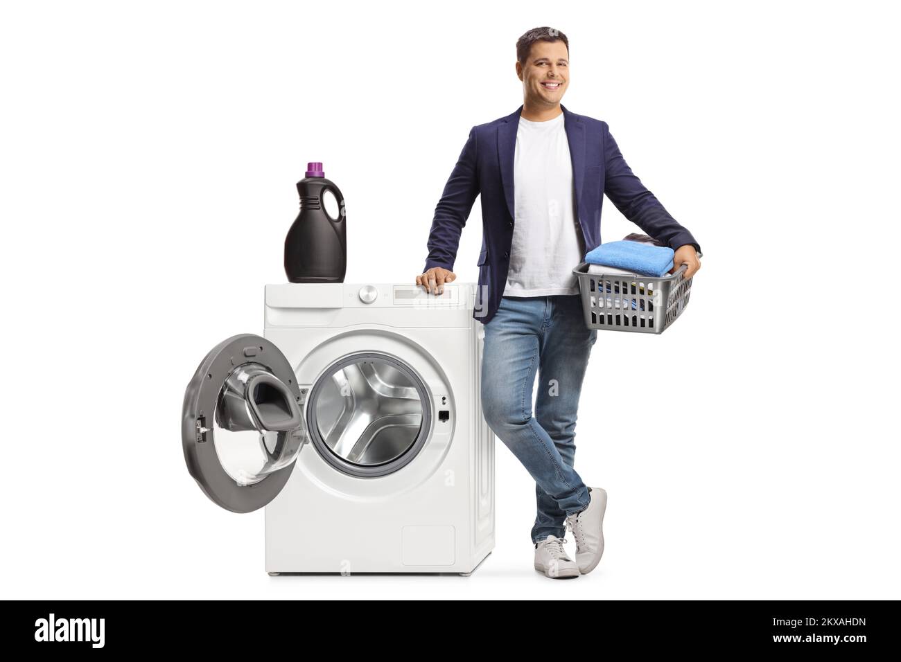 Ein junger Mann lehnt sich an eine Waschmaschine und hält einen Korb mit isolierter Kleidung auf weißem Hintergrund Stockfoto