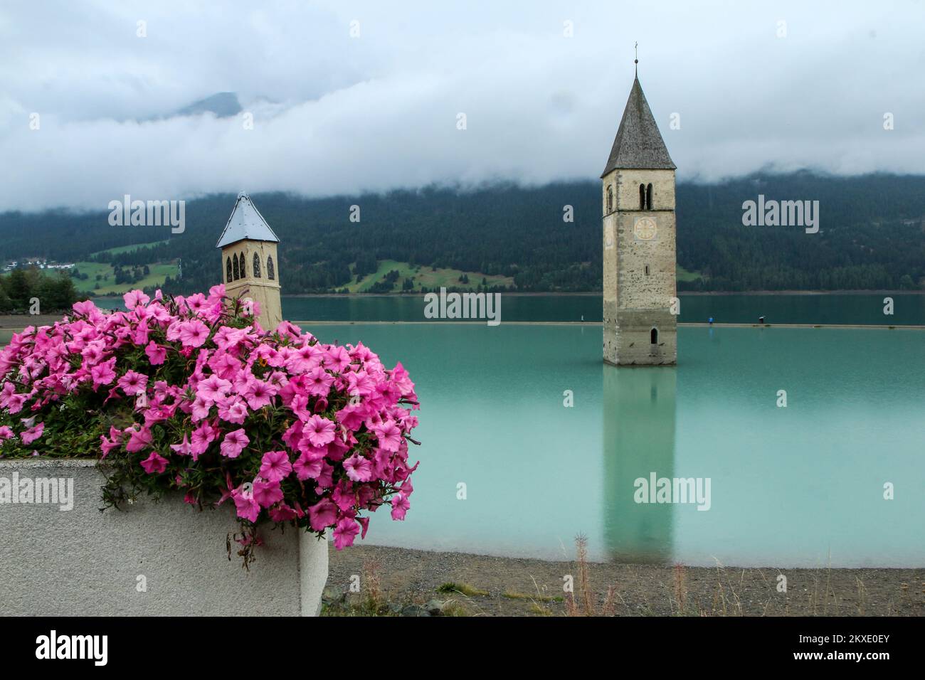 Glockenturm der Katharinenkirche am Lago di Resia mit einer kleinen Miniatur, die in den Blumen steht. Stockfoto