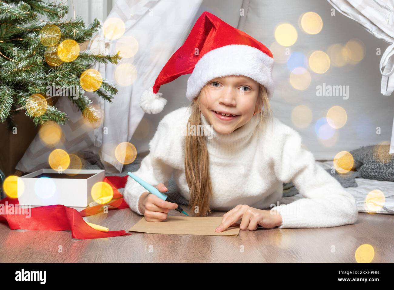 Wunschliste für Weihnachtswunder. Ein lächelndes Mädchen mit Weihnachtsmann-Hut und einem weißen Pullover, das einen Brief schreibt, der Träume für Geschenke an den Weihnachtsmann schreibt. Das Kind liegt zu Hause. Stockfoto