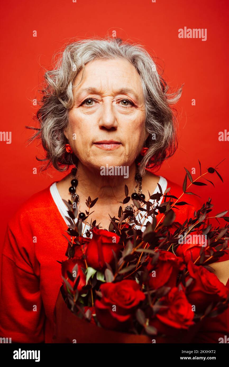 Ältere Frau in roten Kleidern, mit einem roten Rosenstrauß, auf rotem Hintergrund Stockfoto