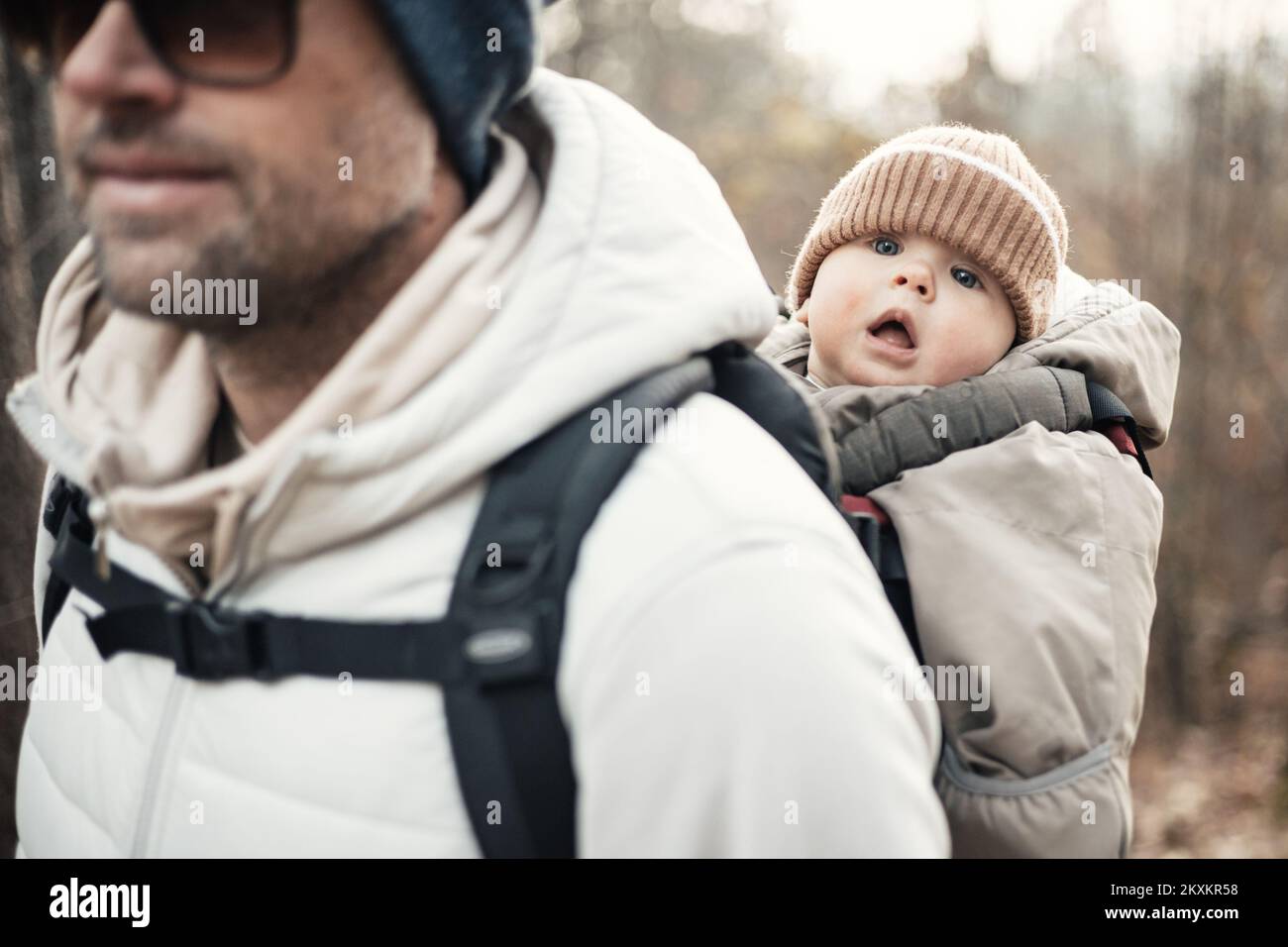 Sportlicher Vater, der seinen kleinen Sohn mit Winteranzug und Mütze im Rucksackträger beim Wandern im Herbstwald trug Stockfoto