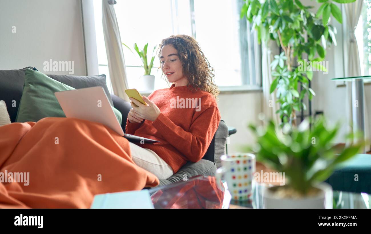 Entspannte junge Frau, die auf dem Sofa sitzt und ein Telefon in einem gemütlichen Wohnzimmer hält. Stockfoto