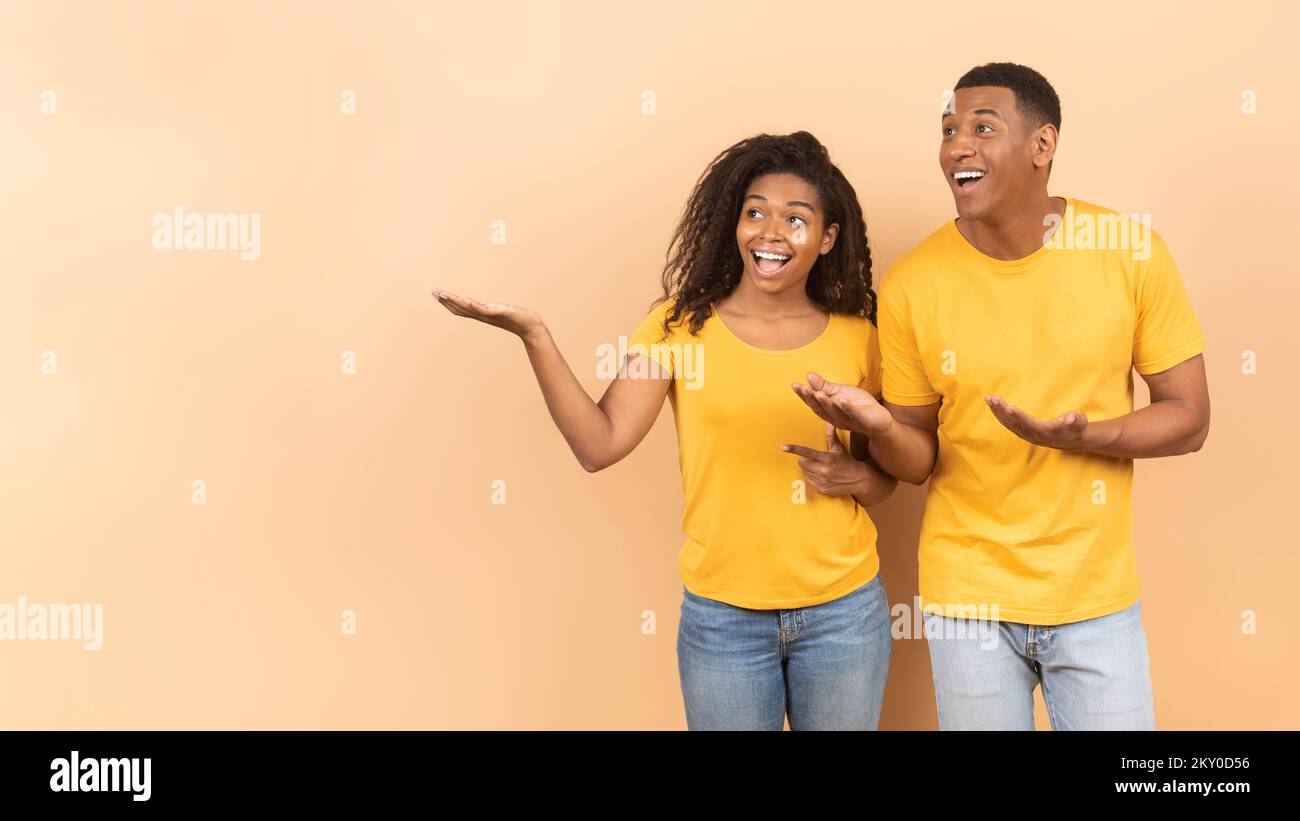 Sieh dir das an. Aufgeregtes afroamerikanisches Paar, das kostenlosen Werbespot vorführt, Pfirsich-Studiohintergrund, Panorama, Banner Stockfoto