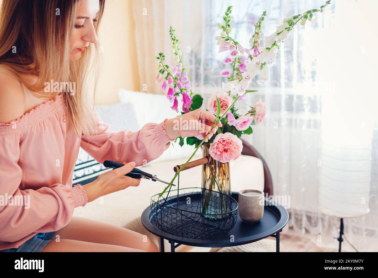 Frau macht Strauß von Rosen und Füchshandschuhen Blumen in Korb in Vase zu Hause gepflückt. Einrichtung und Einrichtung im Sommer. Frische rosa orange Blüten von austin und Stockfoto