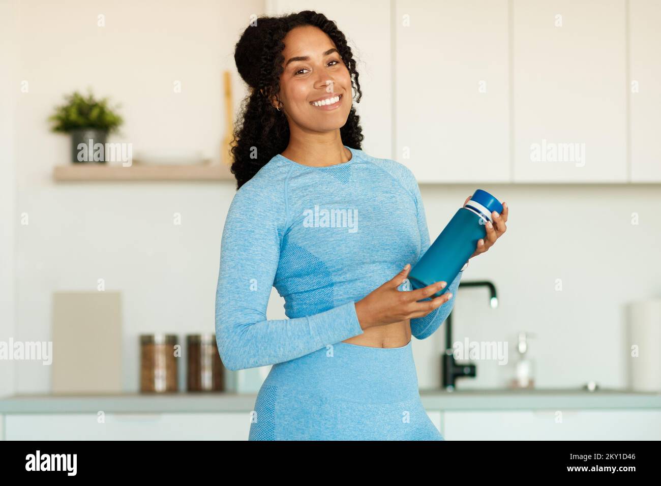 Glückliche junge afroamerikanische Frau mit einer Flasche Protein-Shake in den Händen, die vor der Kamera lächelt und in der Küche steht Stockfoto