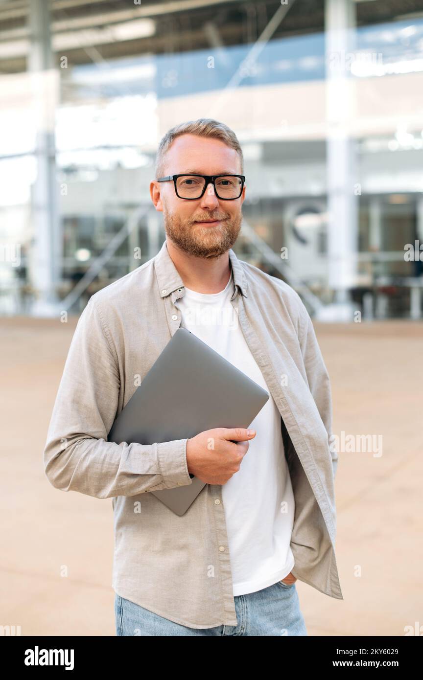 Vertikales Foto eines klugen, selbstbewussten, bärtigen, weißen Mannes mit Brille, in lässiger Kleidung, hält ein Programmierer, seo, einen Laptop in der Hand, sieht in die Kamera und lächelt freundlich Stockfoto