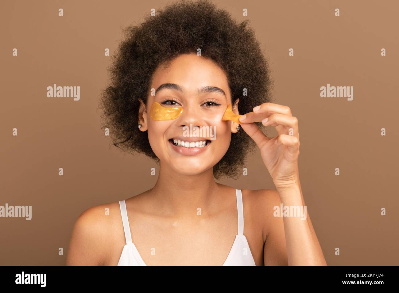 Fröhliche, afroamerikanische Dame mit weißem Oberteil, die Augenflecken aufklebt, isoliert auf braunem Hintergrund Stockfoto