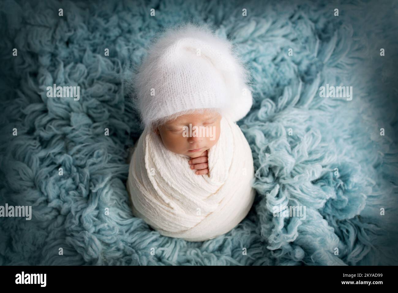 Schlafender neugeborener Junge in den ersten Lebenstagen in einem weißen, weichen Kokon mit einem gestrickten weißen Hut auf blauem Hintergrund Stockfoto