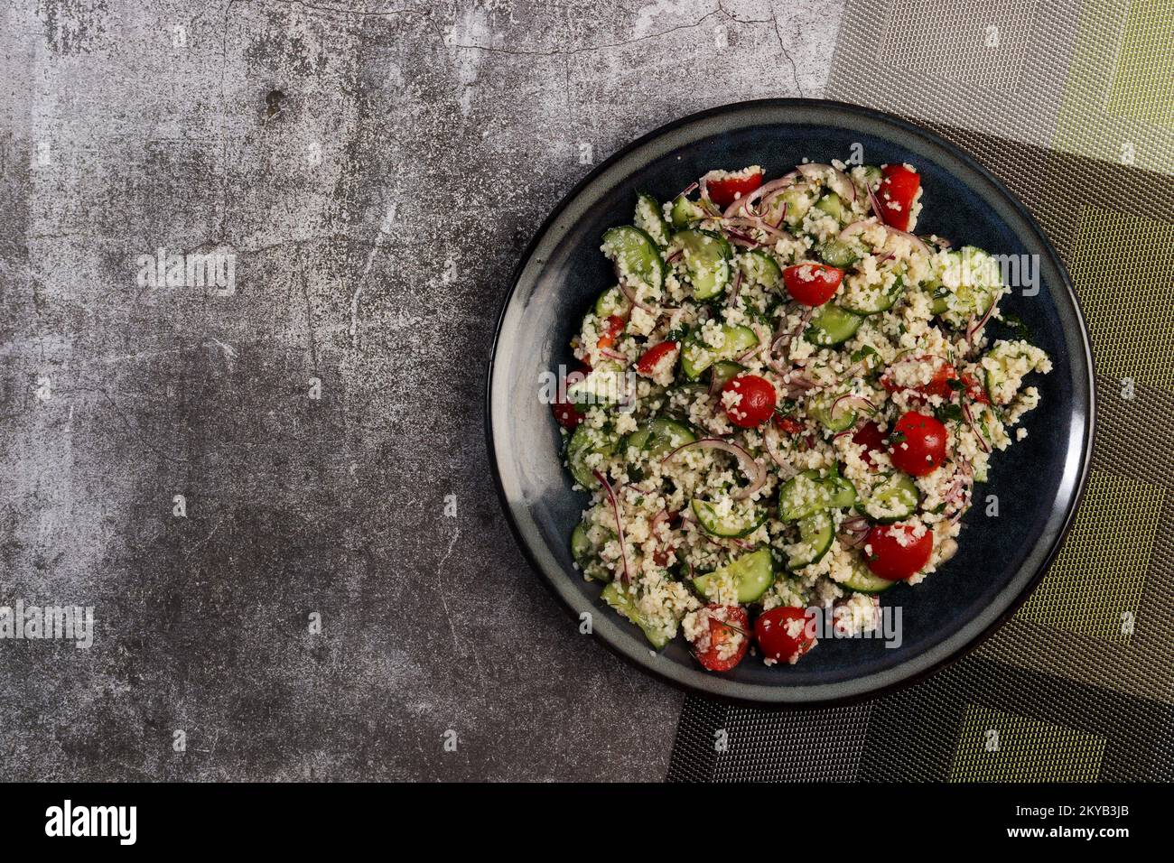 Couscous-Salat mit Gurken, Tomaten und rosafarbenen Zwiebeln auf einem runden Teller mit dunklem Hintergrund. Draufsicht, flach liegend Stockfoto