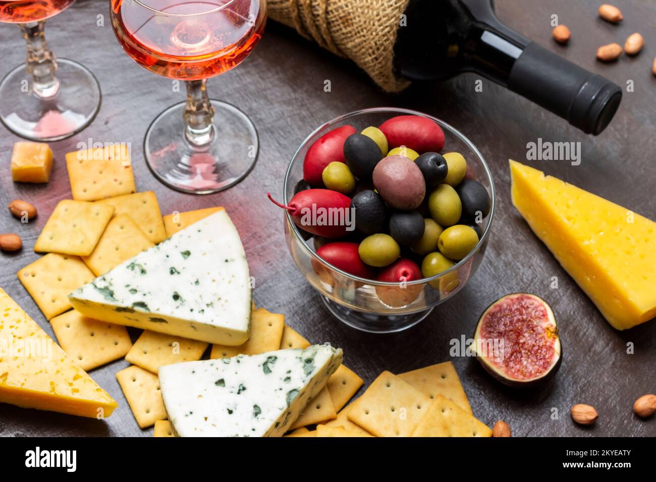 Mehrfarbige Oliven in einer Glasschüssel. Käse und Kekse und eine halbe Feige. Eine Flasche Wein und zwei Gläser Rotwein. Draufsicht. Brauner Hintergrund. Stockfoto