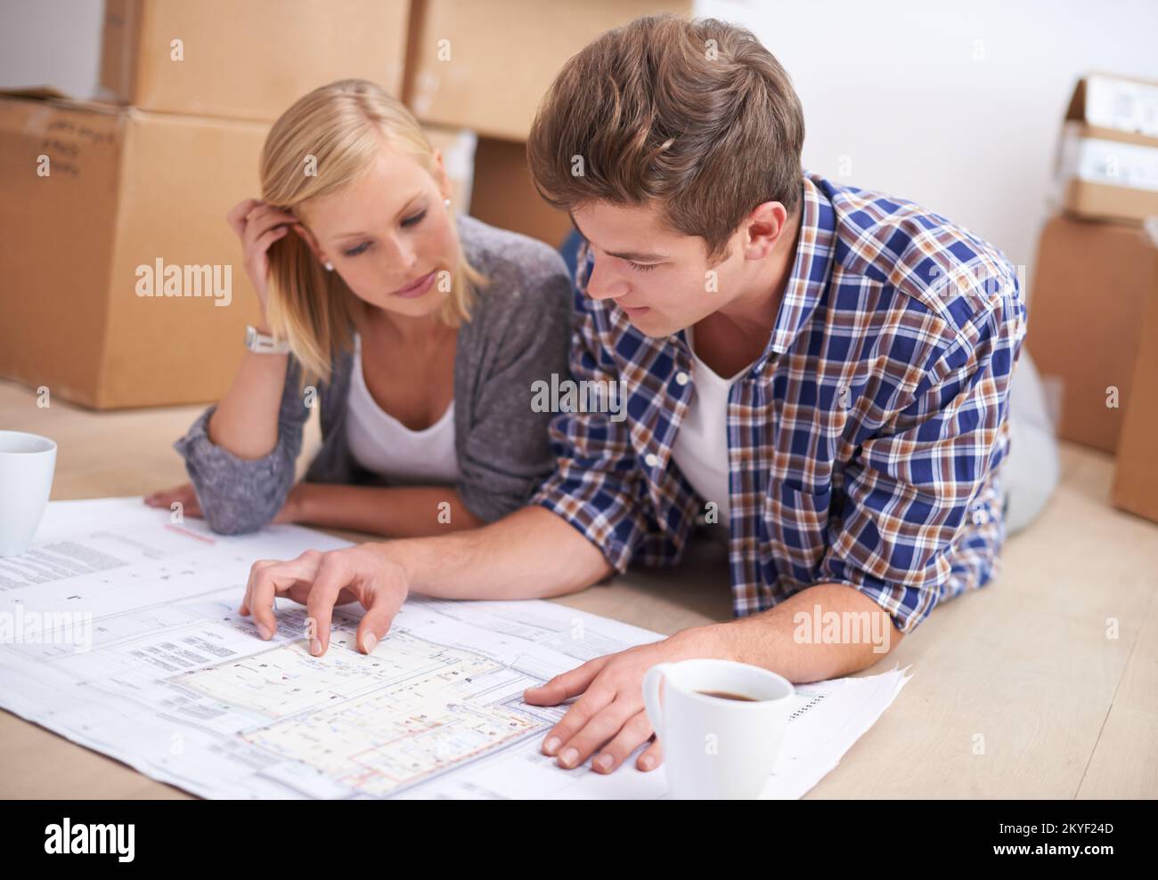 Sie planen ihre Möbel, bevor sie auspacken. Ein junges Paar lag auf dem Boden mit den Bauplänen ihrer neuen Wohnung und Kisten dahinter. Stockfoto