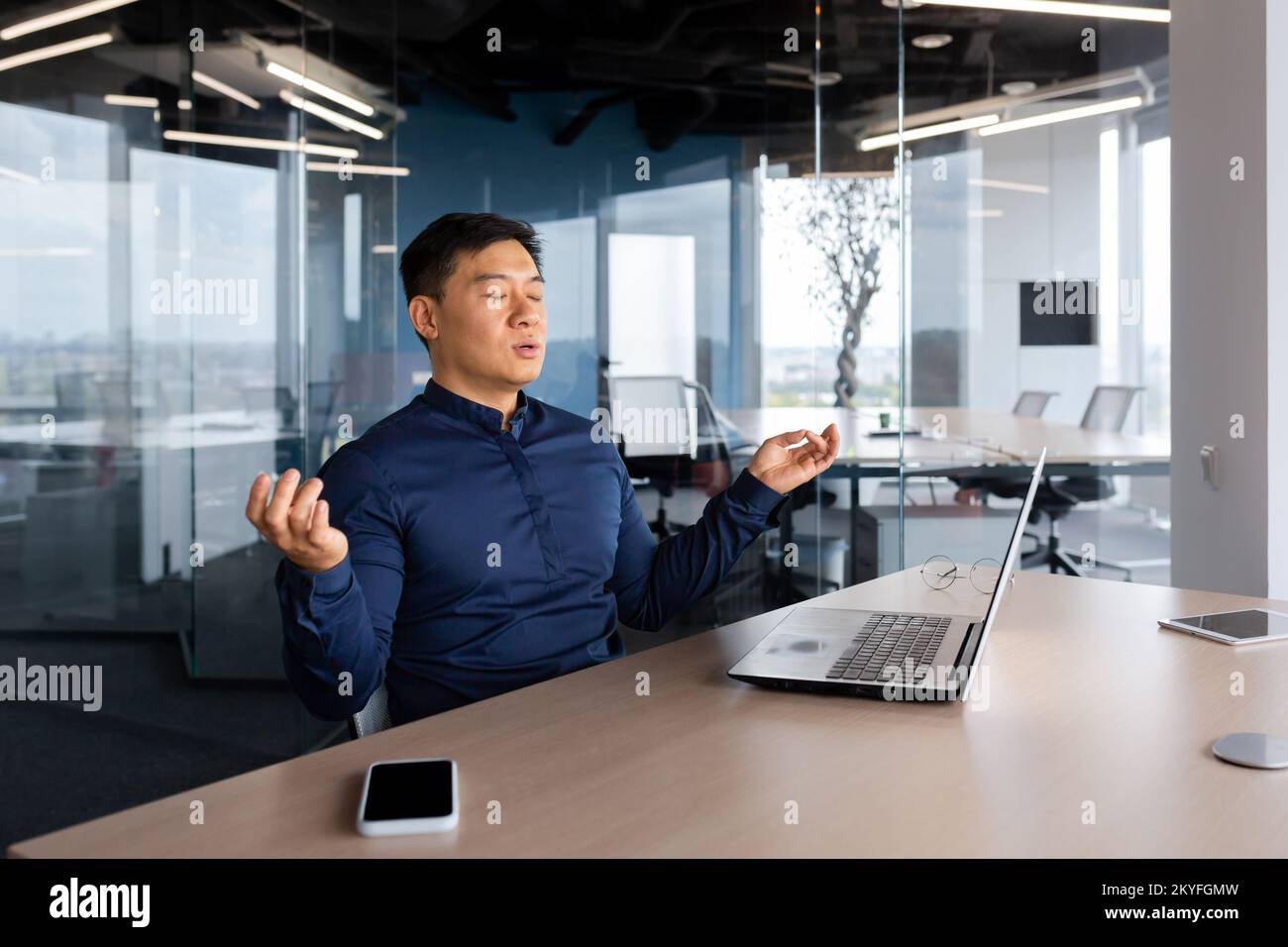 Asiatischer Geschäftsmann, der im Büro meditiert, Mann, der am Schreibtisch mit einem Laptop arbeitet, Arbeiter mit geschlossenen Augen im Lotus posieren im Gebäude. Stockfoto