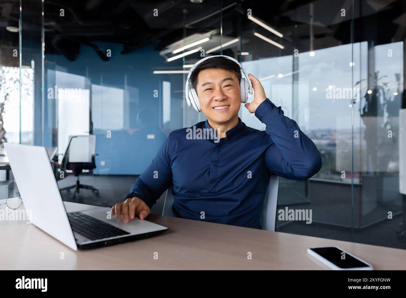 Porträt eines glücklichen asiatischen Arbeiters in einem modernen Büro, eines Mannes, der in die Kamera schaut und lächelt, eines Chefprogrammierers im Hemd, der Kopfhörer verwendet, um sich Videos anzusehen und Musik bei der Arbeit zu hören. Stockfoto