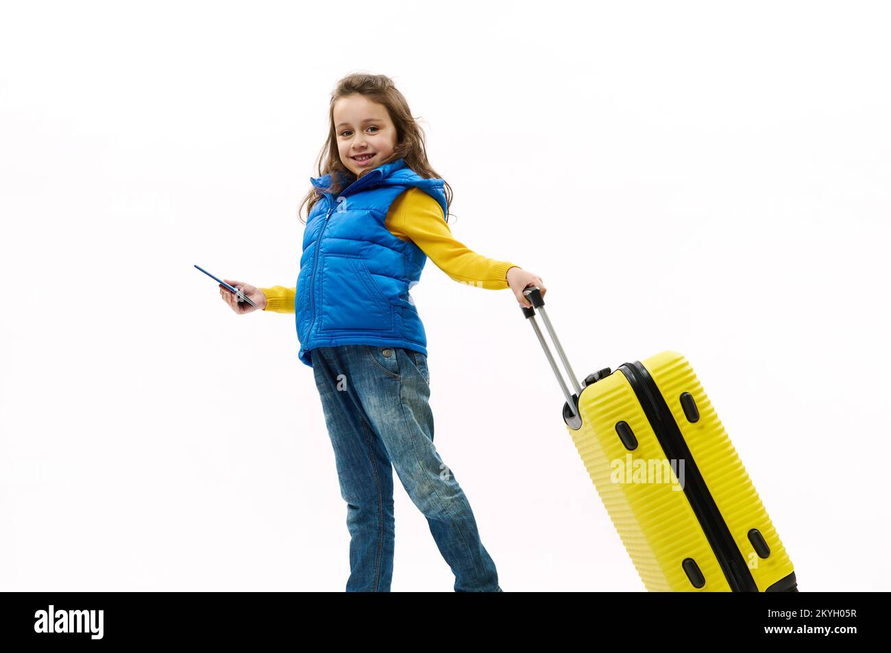 Stilvolles Mädchen für Kinder in blauen Jeans, gelbem Hemd und Daunenjacke, mit Koffer und Bordkarte, Reisen ins Ausland Stockfoto