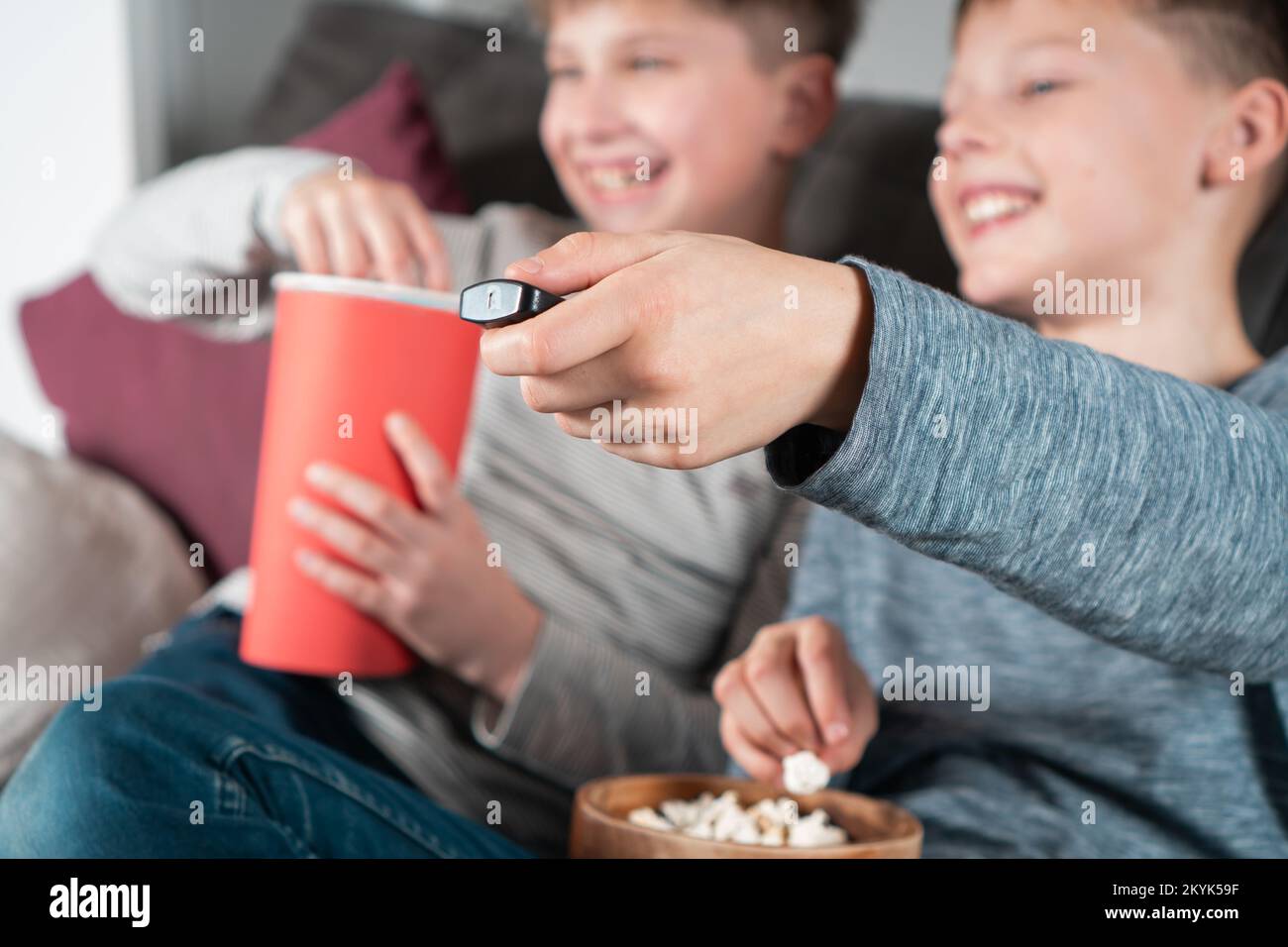 Fröhliche Jungs im Schulalter sitzen auf dem Sofa, essen Popcorn und schauen fern. Konzentrieren Sie sich auf die ausgestreckte Hand mit der TV-Fernbedienung. Die Kinder haben Spaß im Zimmer Stockfoto