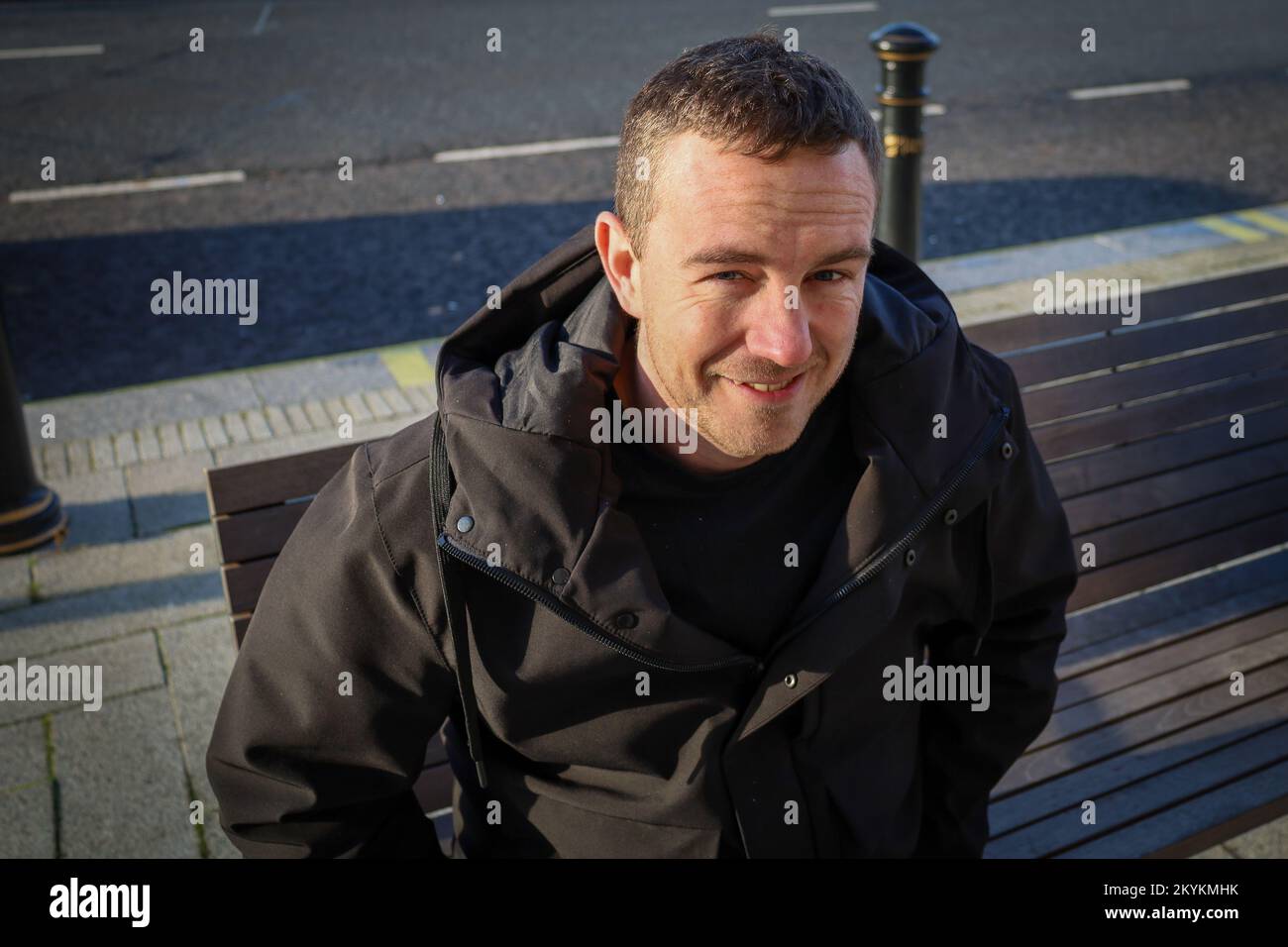 Ein Mann, der auf einer Bank in Liverpool sitzt und in die Kamera lächelt Stockfoto