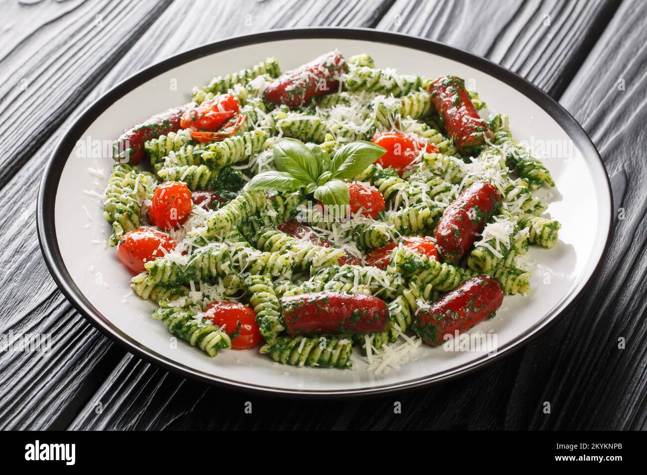 Herzhaftes Mittagessen Fusilli Pasta mit Würstchen, Tomaten, grüner Pesto-Sauce und Käse aus nächster Nähe auf einem Teller auf dem Tisch. Horizontal Stockfoto