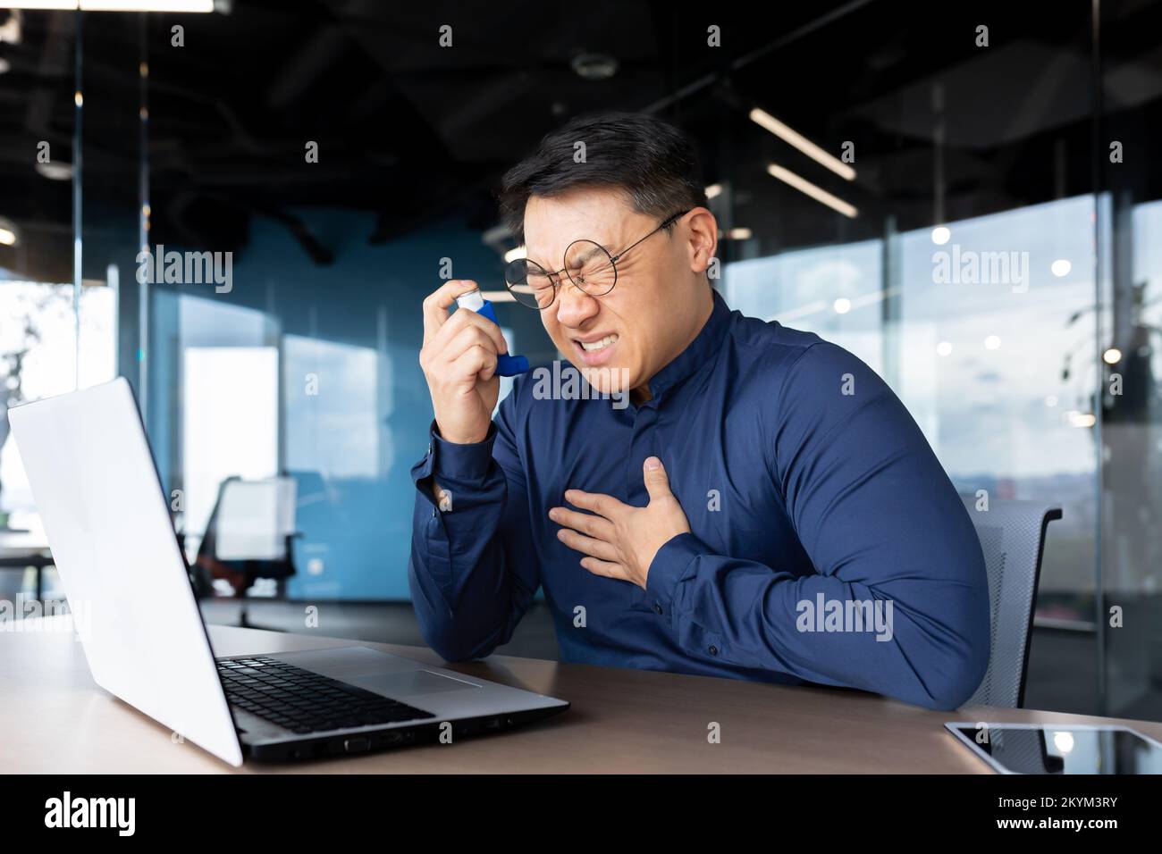 Asthmaanfall bei der Arbeit, asiatischer Geschäftsmann mit Atembeschwerden, Mann mit Inhalator und Medikamenten zur Erleichterung der Atmung, Mitarbeiter im Büro mit Laptop bei der Arbeit. Stockfoto
