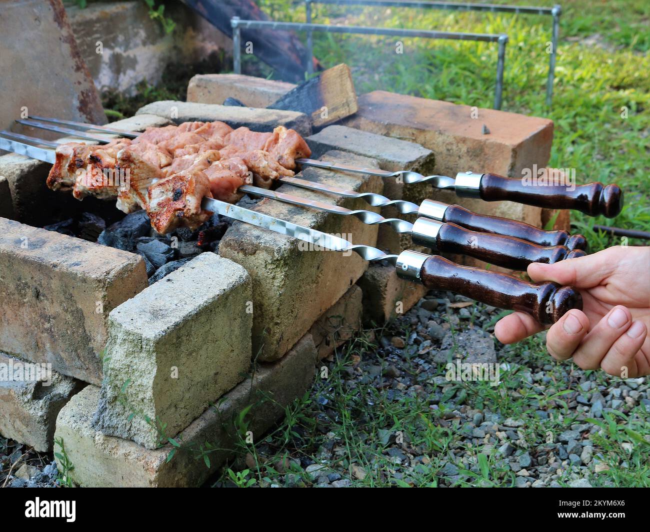 Ein willkürlicher Ort aus Ziegelsteinen für ein Feuer und Grillen marinierter Schweinespieße, Kochen von Fleisch auf Spießen mit wunderschönen Holzgriffen auf Kohlen im Freien Stockfoto