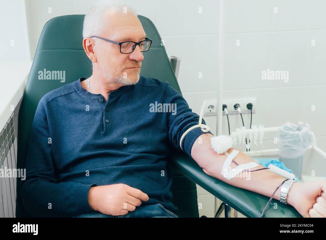 Spender spendet Blut. 50-60 Jahre alter Mann mit Brille sitzt in einem Stuhl in der medizinischen Klinik und spendet Blut aus der Vene. Authentische Szene. Freiwilliger spendet Blut, um Leben zu retten. Stockfoto