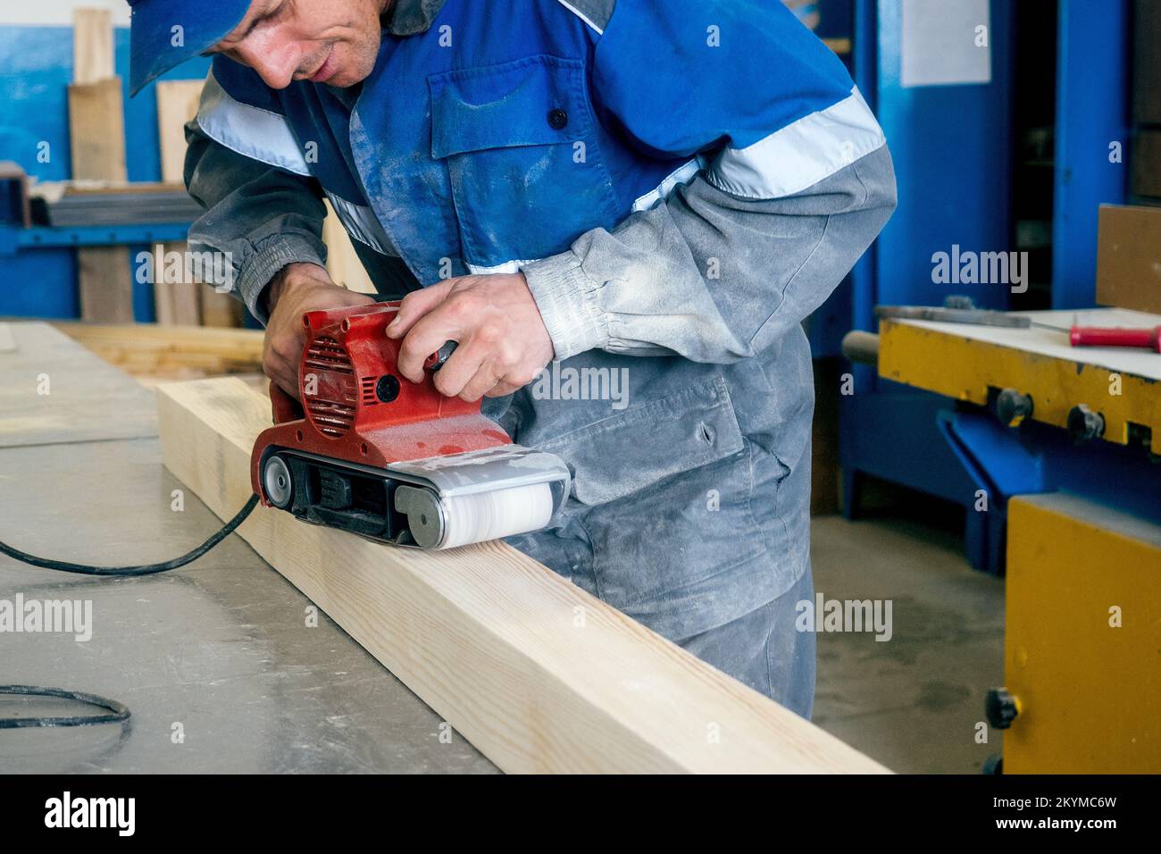 Professioneller Holzarbeiter schleift Holzstange auf Werkbank in Zimmereiwerkstatt mit Werkzeug. Arbeiter mittleren Alters in Overalls verarbeiten Holz mit einem elektrischen Schleifer. Echter Workflow... Stockfoto
