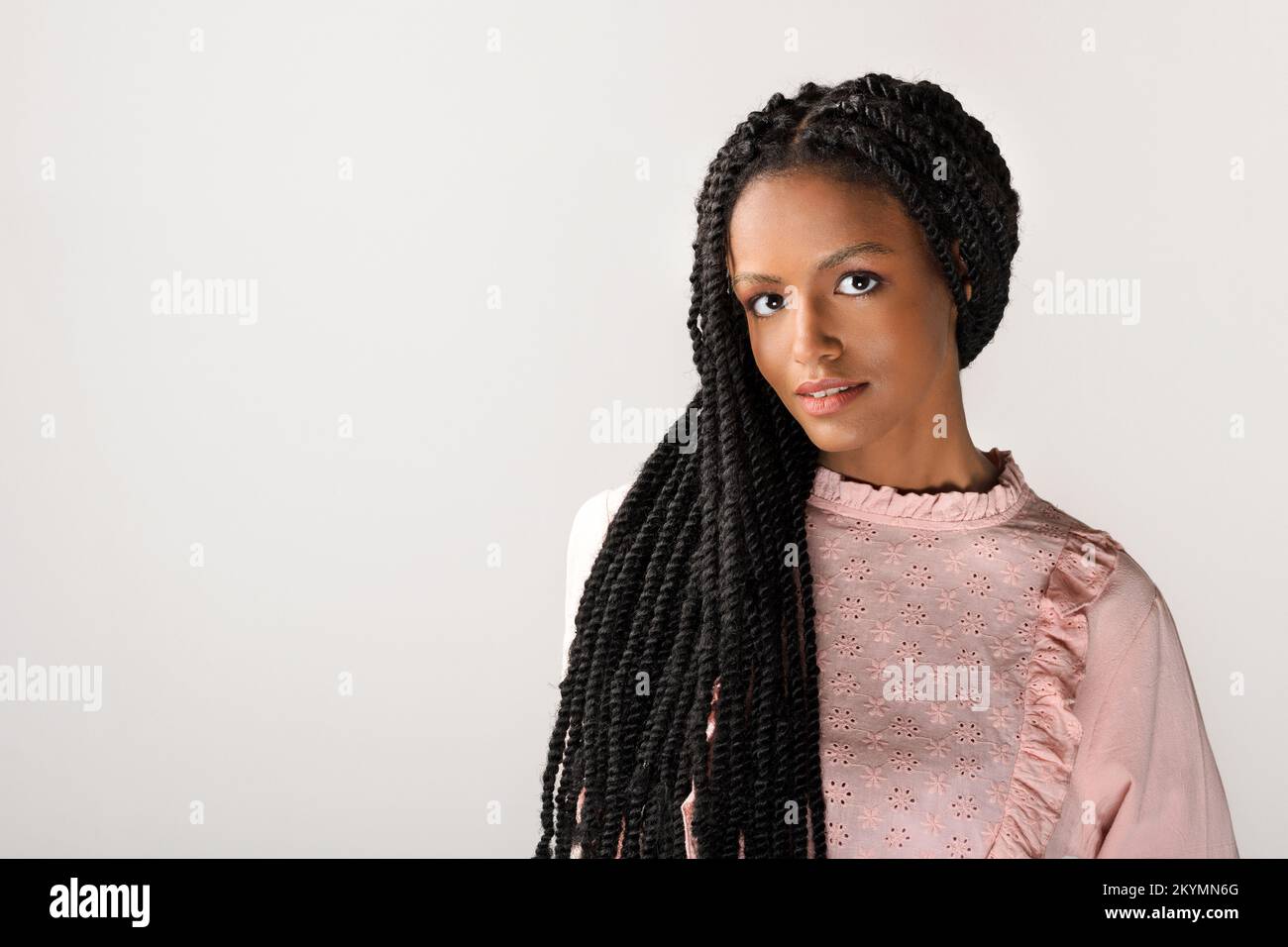 Sanfte junge afroamerikanische Frau in bescheidenem rosa Kleid, die Haare in stilvollen langen Afro-Geflechten hat und auf grauen Hintergrund auf die Kamera schaut Stockfoto