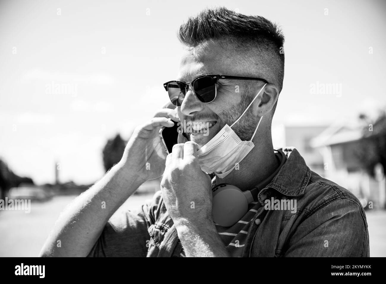 Lächelnder Mann mit Kopfhörern um den Hals und tiefer gefachte OP-Maske nach Ausbruch des Coronavirus reden - Junge Mann im Gespräch mit Handy-Outdoo Stockfoto