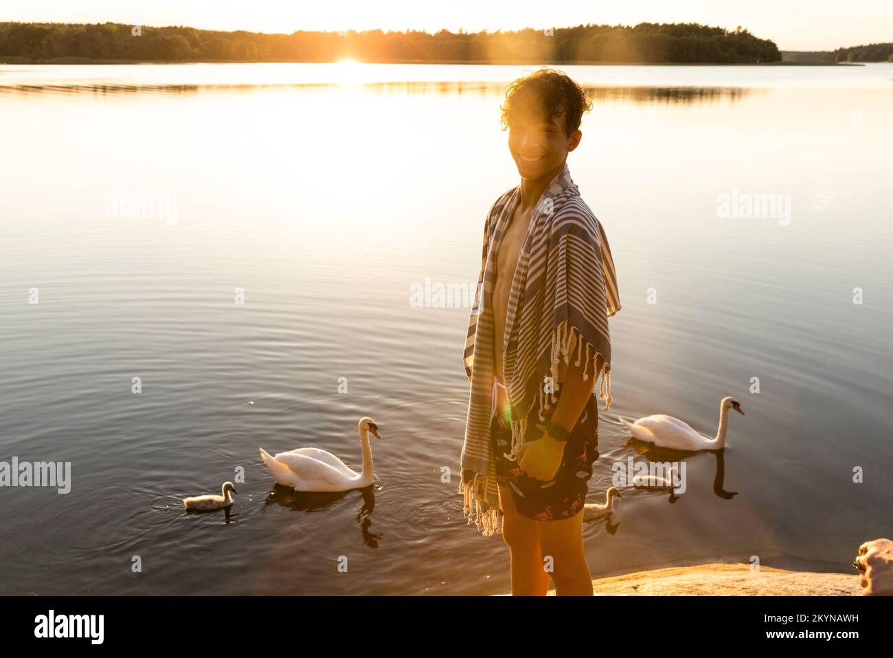 Porträt eines glücklichen Jungen, der in der Nähe des Sees steht und im Hintergrund Schwäne schwimmt Stockfoto