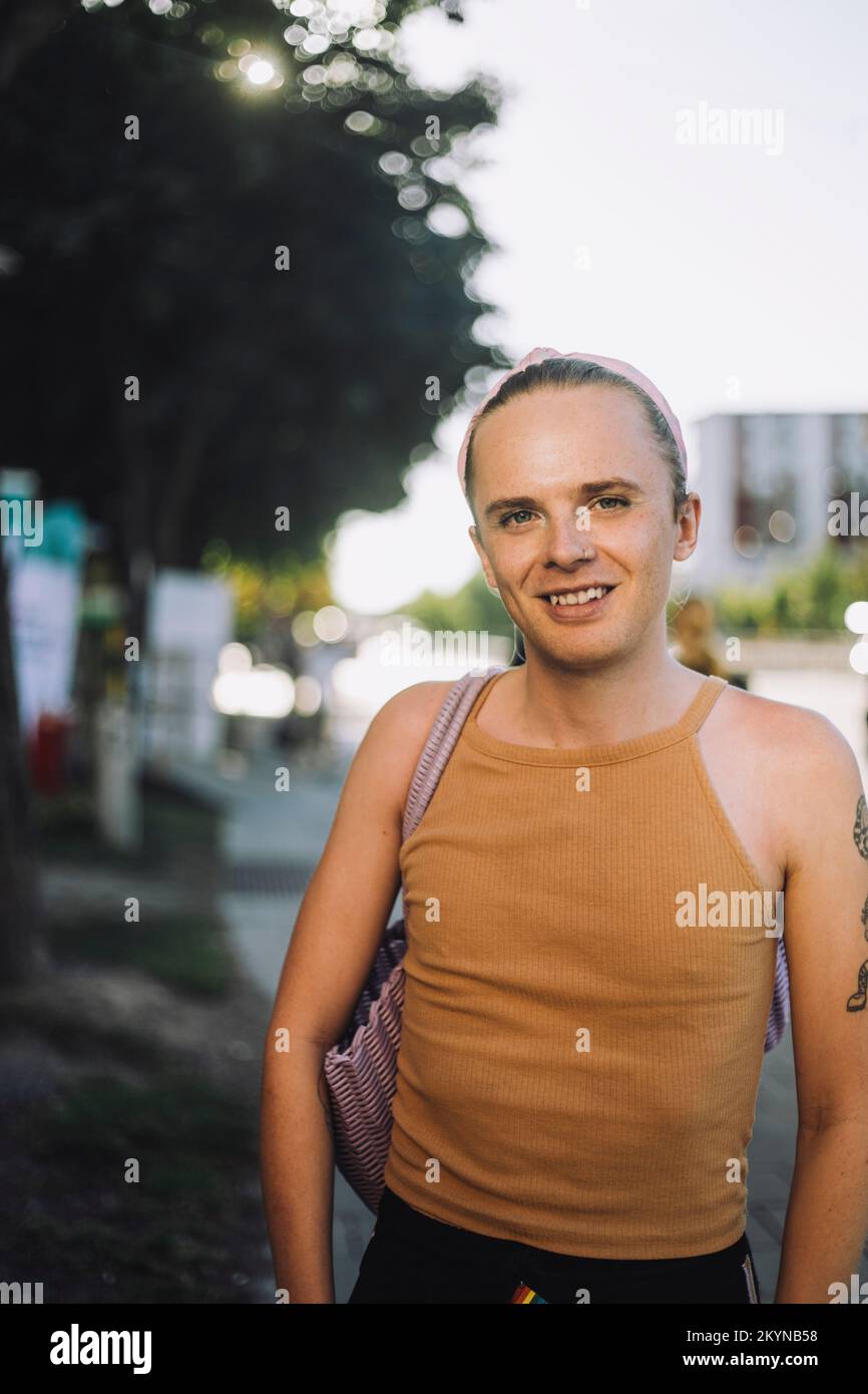 Porträt einer lächelnden nicht-binären Person im Stehen mit Schultertasche Stockfoto