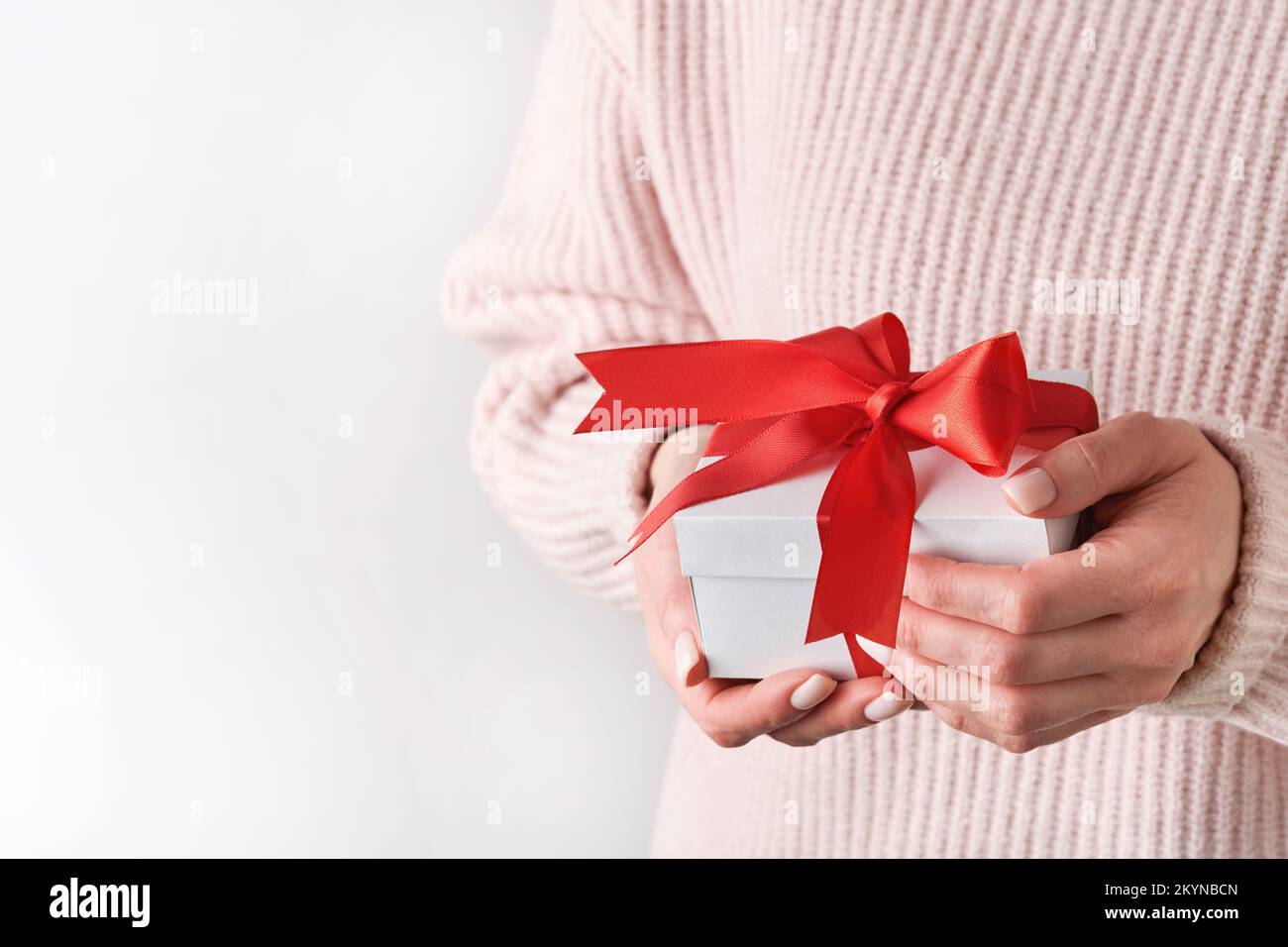 Weibliche Hände in pinkfarbenem Wollpullover mit weißer Geschenkbox mit rotem Band auf hellem Hintergrund. Weihnachtskomposition, Geburtstagskomposition oder Hochzeitskomposition mit Exemplaren Stockfoto