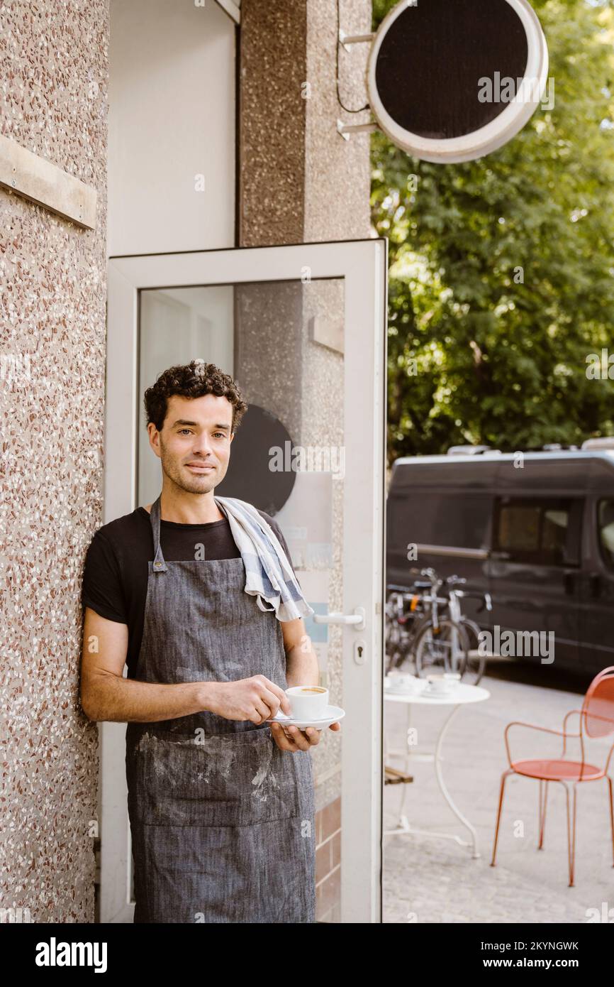 Ein männlicher Cafe-Besitzer, der eine Schürze trägt und die Kaffeetasse an die Wand lehnt Stockfoto