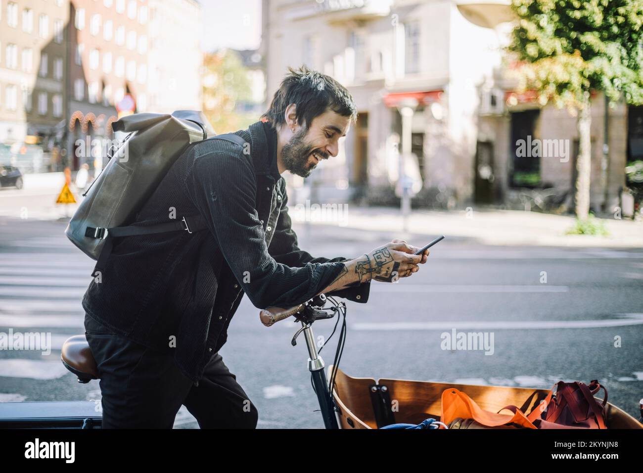 Seitenansicht eines lächelnden Geschäftsmanns, der ein Smartphone benutzt, während er sich auf ein Cargo Bike lehnt Stockfoto