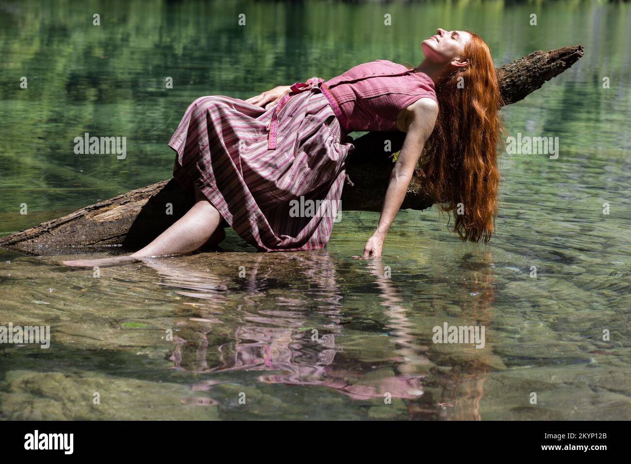 Eine junge Frau liegt mit der Hand auf einem Baumstamm in einem smaragdfarbenen Bergsee Stockfoto