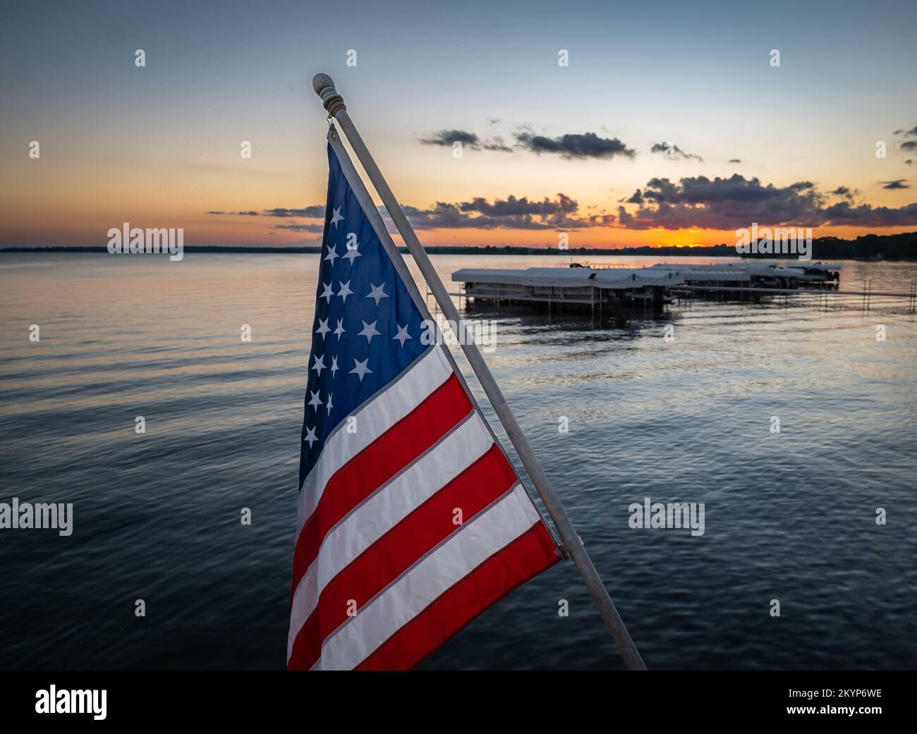 Amerikanische Flagge an einem angewinkelten Stab mit Booten an einem Seehafen und Wasser nach Sonnenuntergang in der Ferne in der Dämmerung. Stockfoto