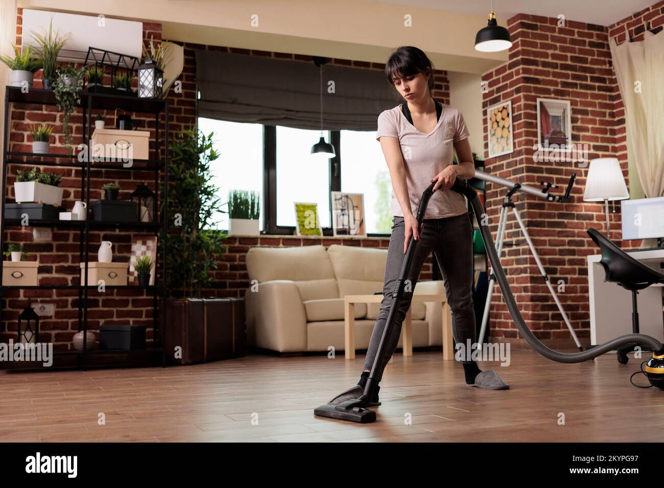 Die Ehefrau kümmert sich um den Frühjahrsputz, saugt den Staub im Wohnzimmer zu Hause ab. Hausfrau in Hausschuhen macht Hausarbeiten mit Hingabe, räumt auf und putzt alles. Stockfoto