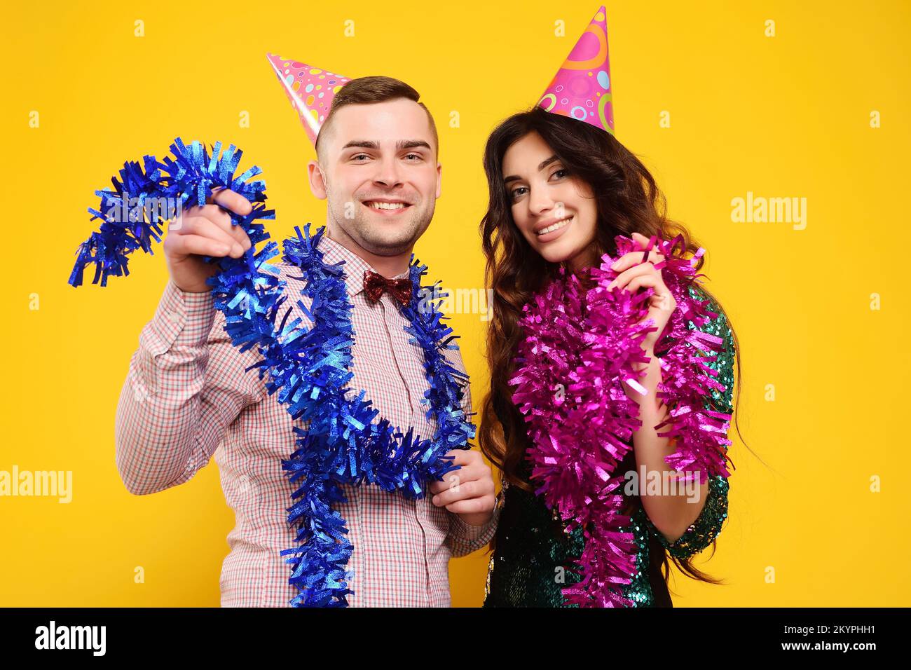 Mann und Frau in Partymütze und mit pinkfarbener und blauer Boa um den Hals, die auf gelbem Hintergrund lächelt Stockfoto